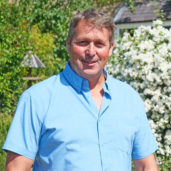 Man in a light blue shirt smiles outdoors, set against greenery and white flowering bushes.
