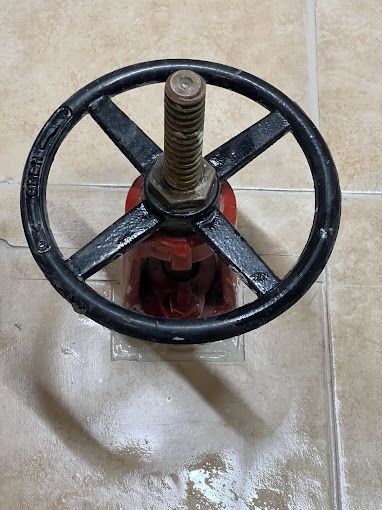 A red valve with a black steering wheel is sitting on a tiled floor.
