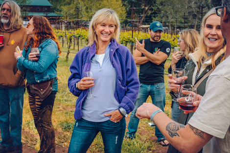 Group of people at a vineyard, some holding wine glasses. Woman in purple jacket smiles, looking at the camera.