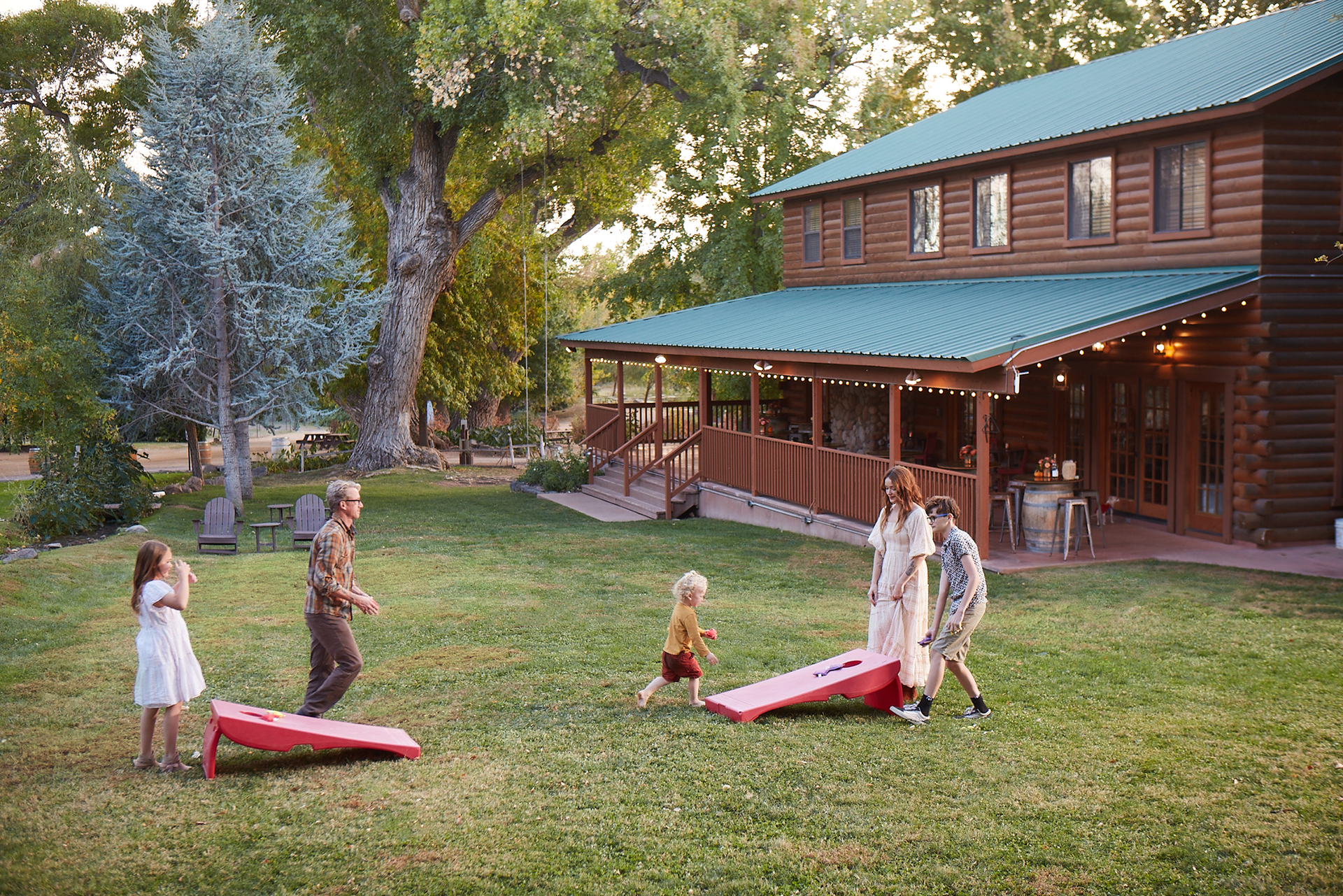Family playing cornhole in a yard with a log cabin house.