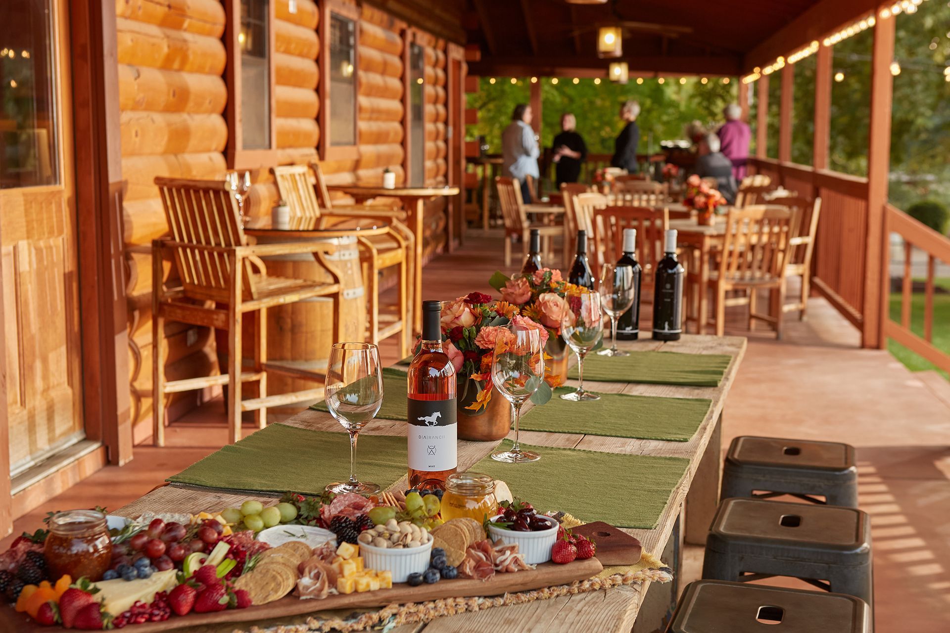 Outdoor dining setup with wine, charcuterie, and people on a porch of a log cabin.