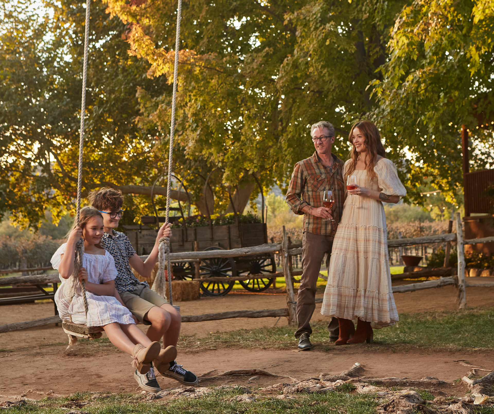 Family enjoys a sunny day outdoors. A young girl and boy swing. A woman and man watch nearby.