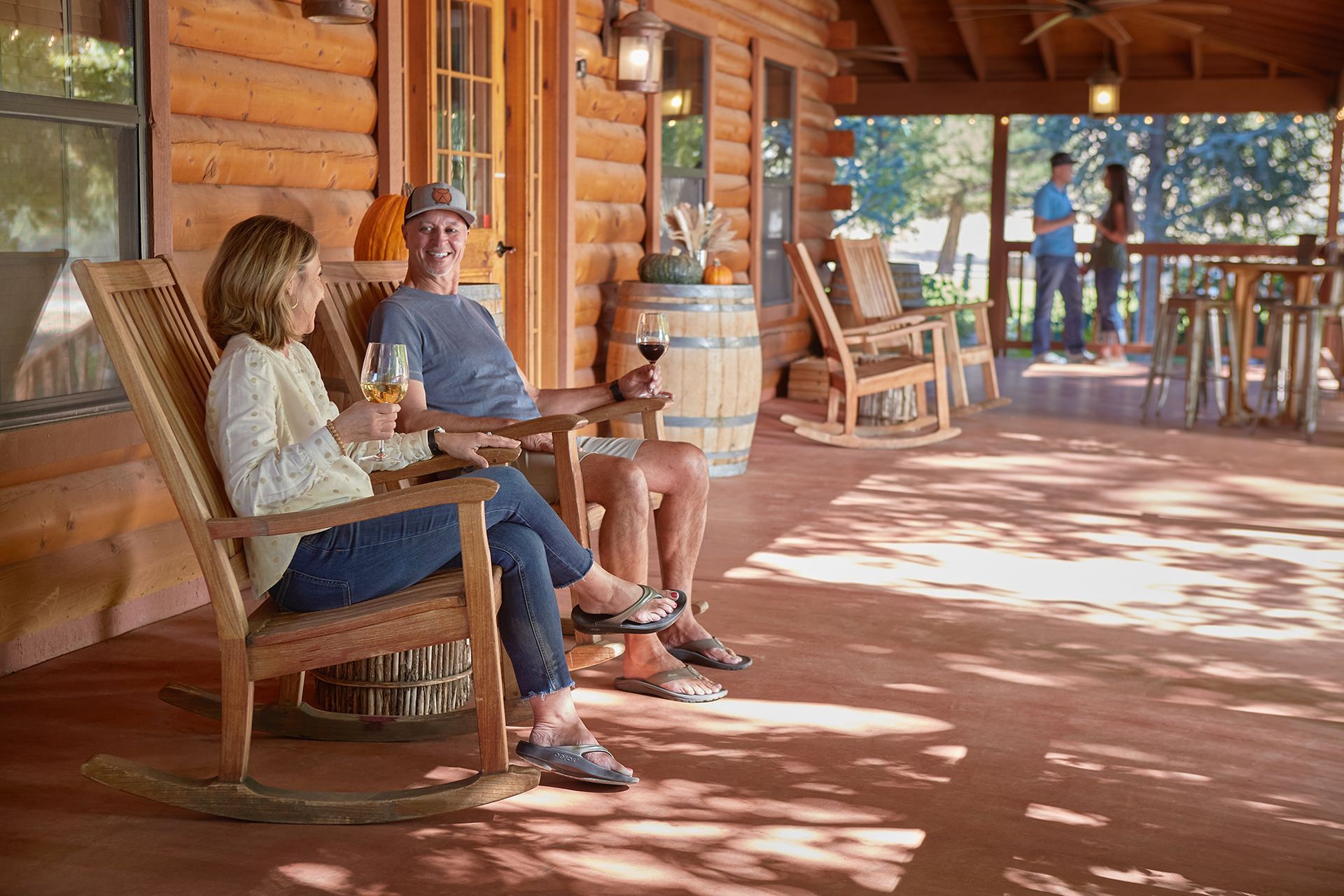 Couple relaxing on a wooden porch, drinking wine, in front of a log cabin; other people in the background.