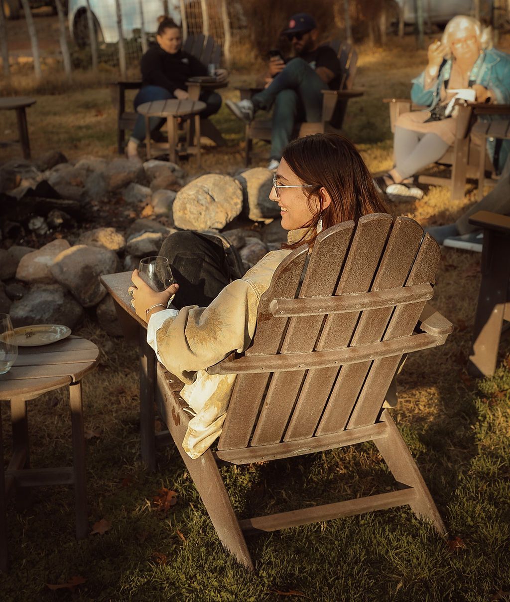 Woman in glasses relaxes in wooden chair outdoors, surrounded by others.