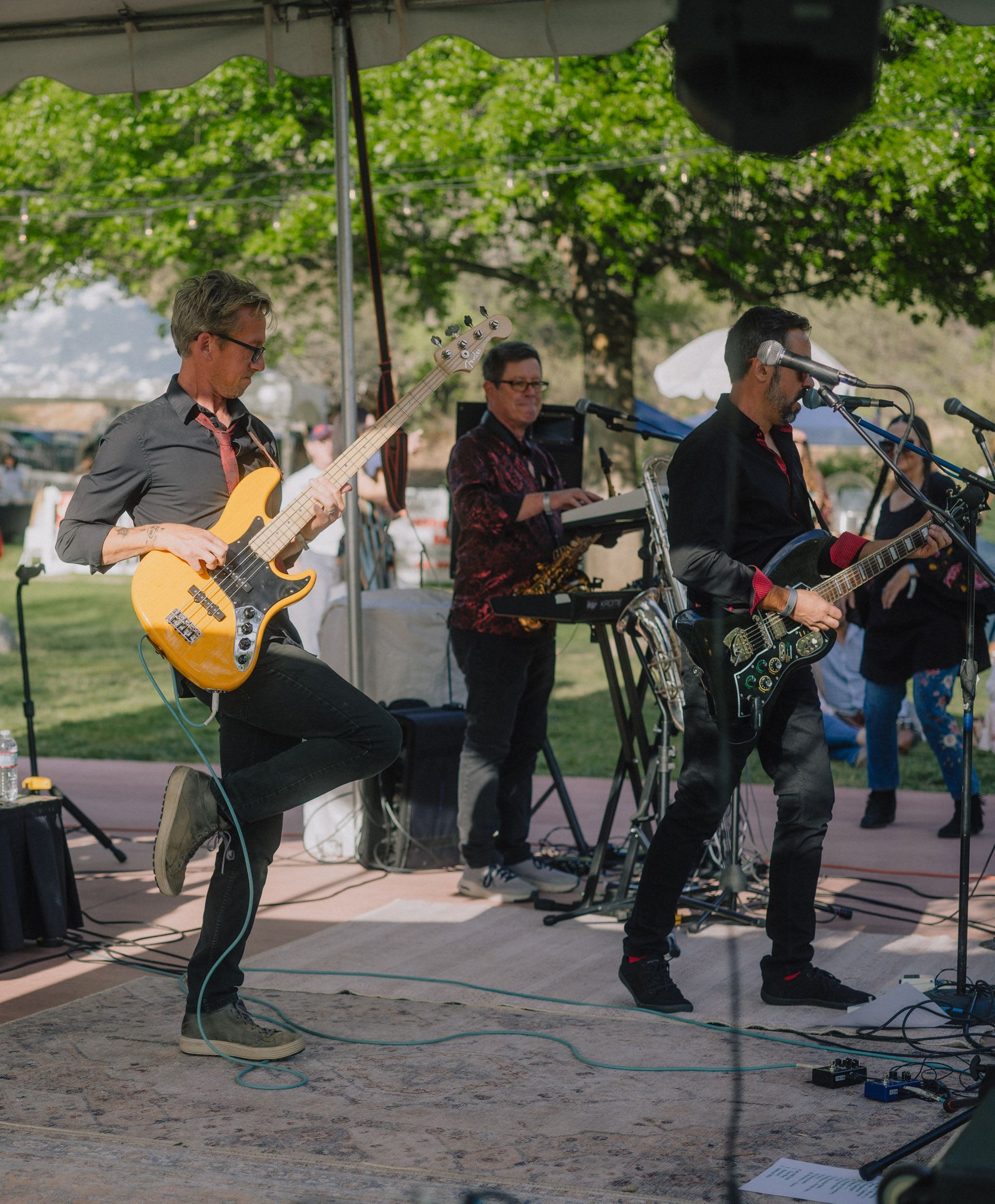 A band plays outdoors: bassist with raised leg, guitarist, keyboardist, drummer, and vocalist.