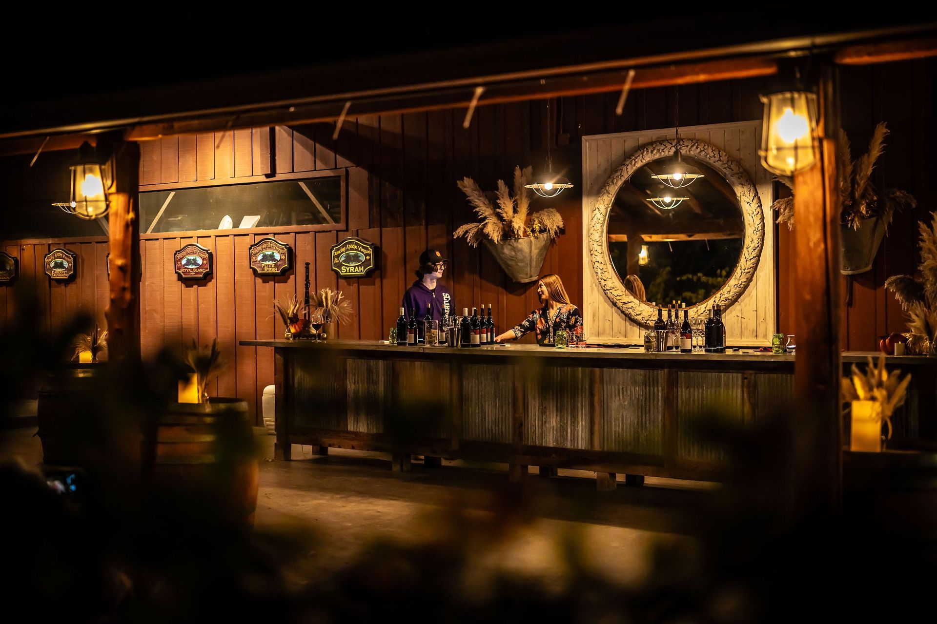 Wooden bar at night with two bartenders, ornate mirror, and lanterns.