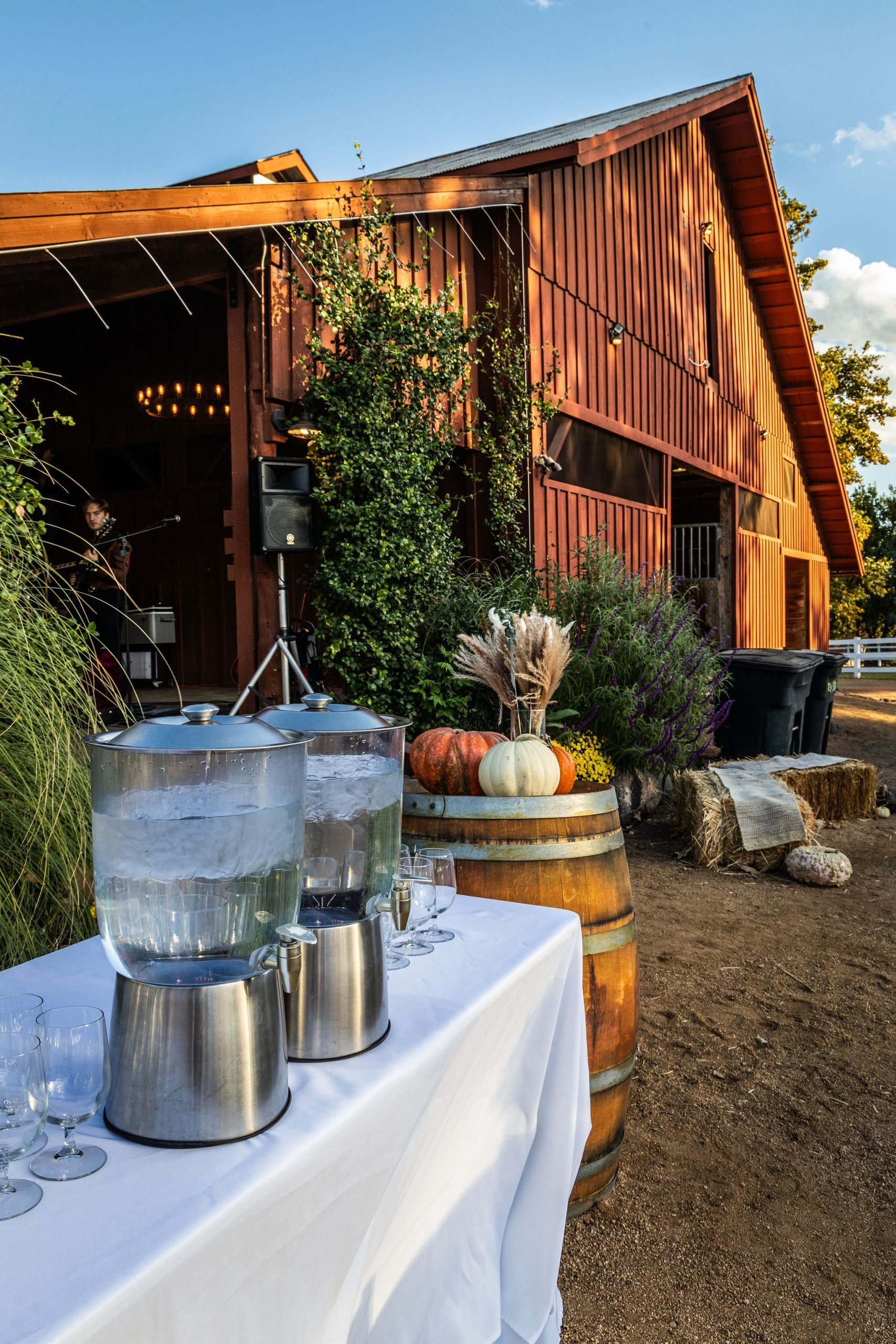 Two water dispensers on a table with a white cloth in front of a barn. Fall decorations.