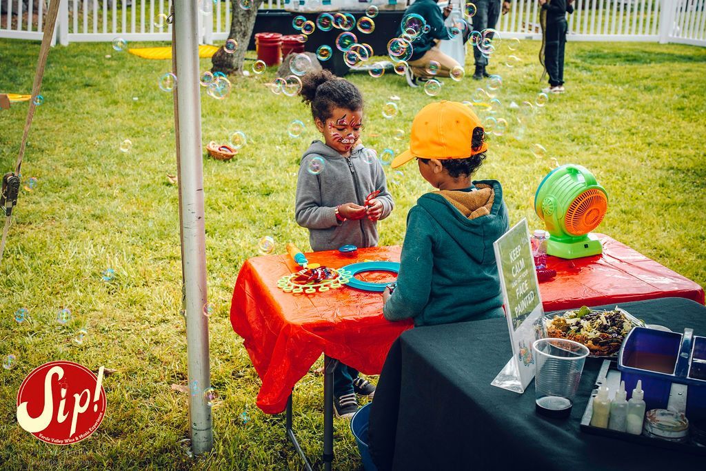 Two children playing at a table outdoors with a bubble machine, grass, and a white fence.