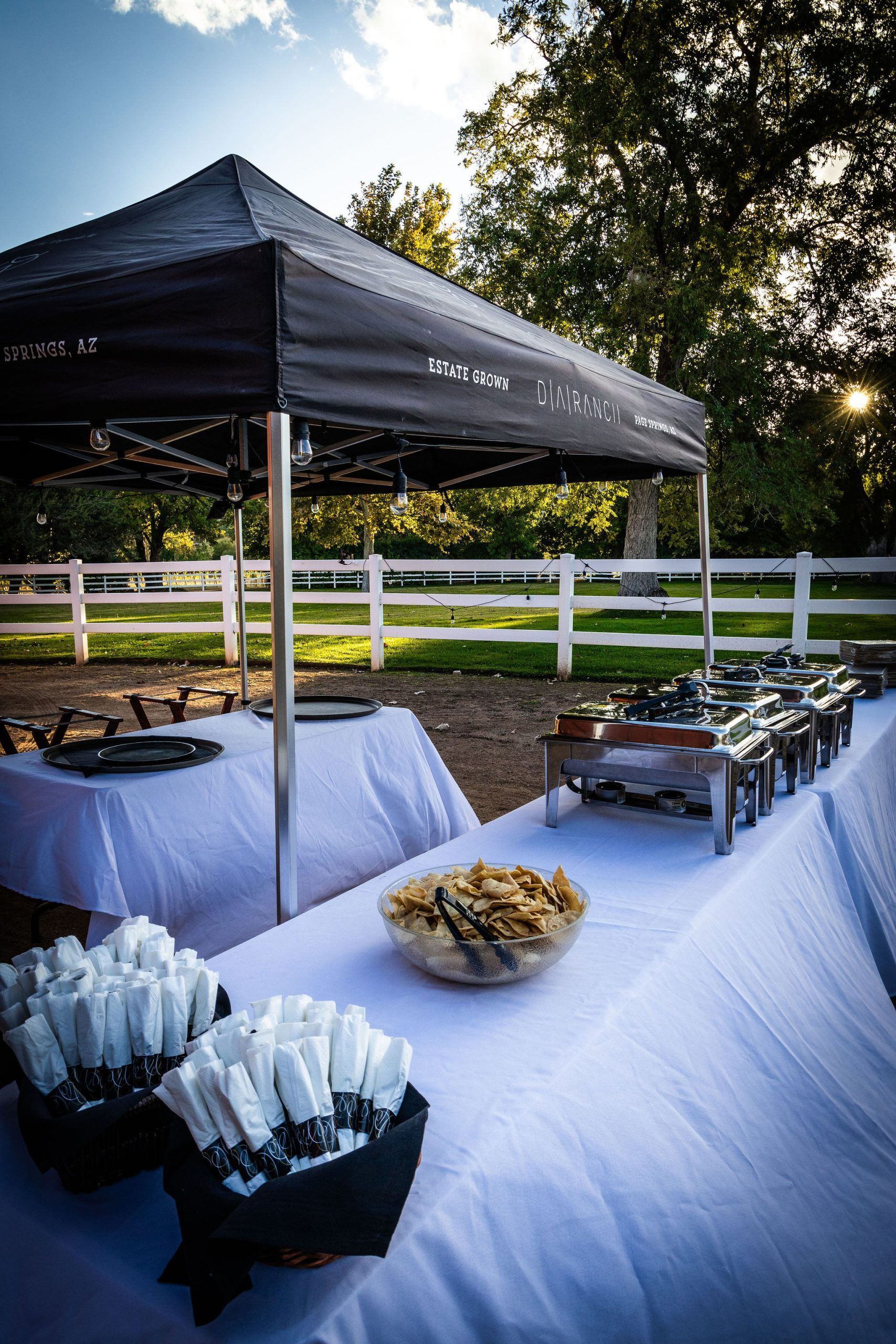 Catered outdoor buffet under a black tent with white tablecloths, food warmers, and snacks, set against a white fence and trees.