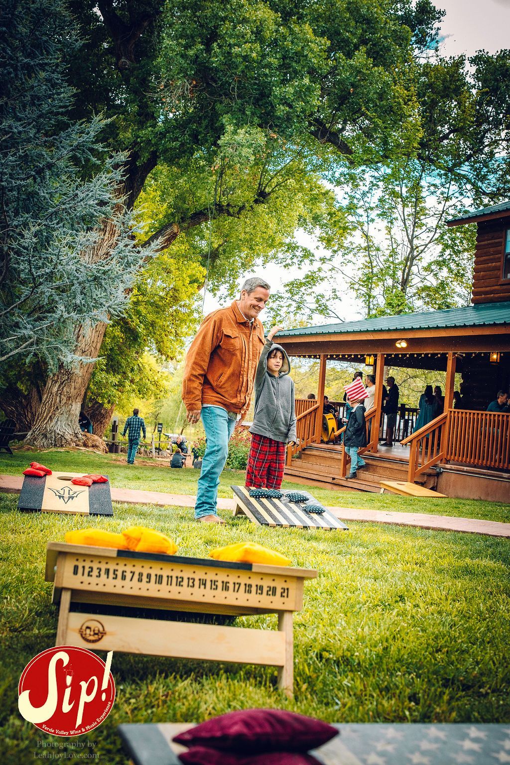 Man and child playing cornhole outside a cabin on a grassy lawn; others are on the porch.