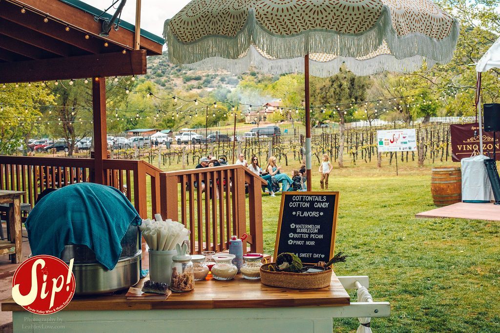 Outdoor bar at a vineyard with drinks, a decorative umbrella, and people in the background.