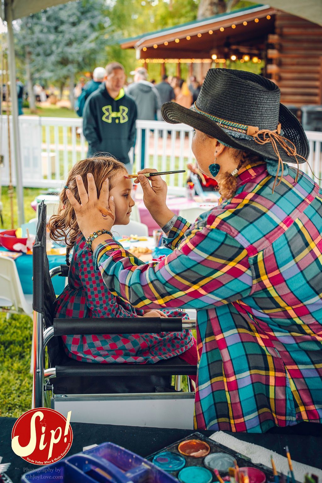 Woman paints a young girl's face outdoors. The girl sits in a chair. Other people and buildings are in the background.