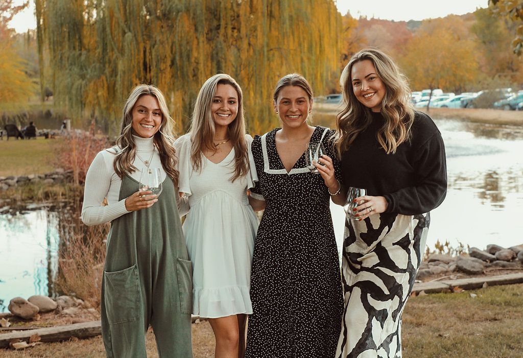 Four women smiling, holding drinks, posing near a pond with a weeping willow in autumn.