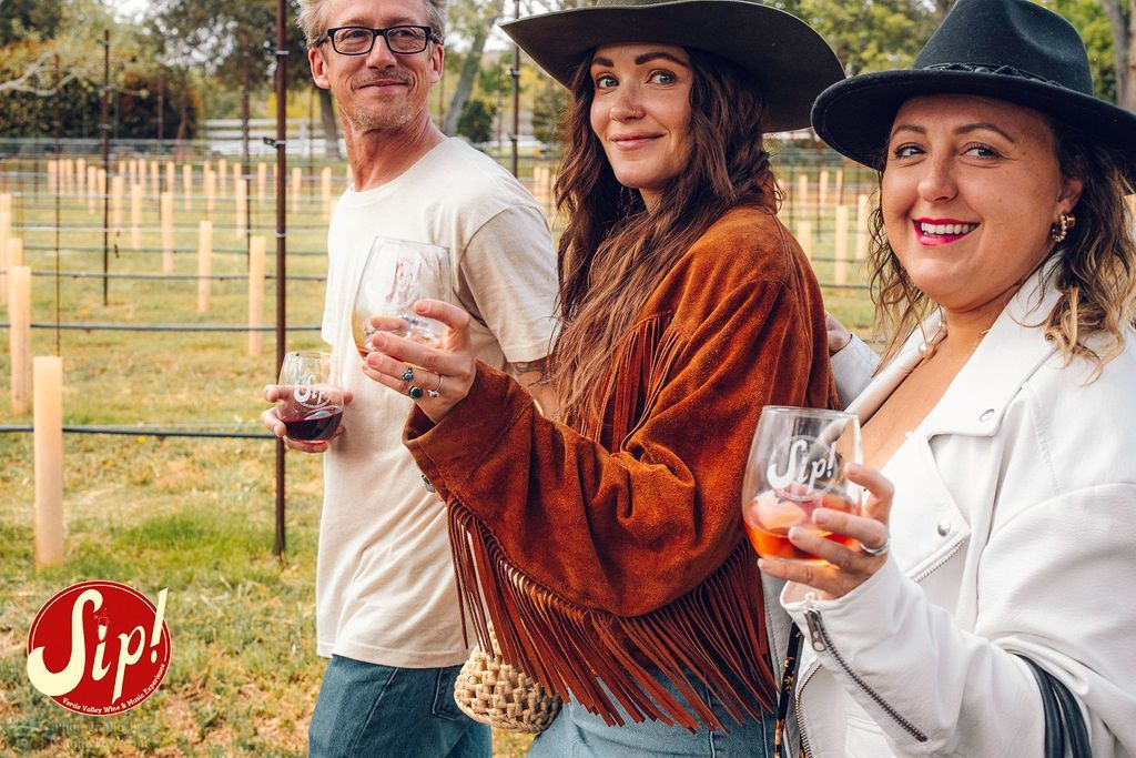 Three people with wine glasses walk through a vineyard. Two women in hats smile.