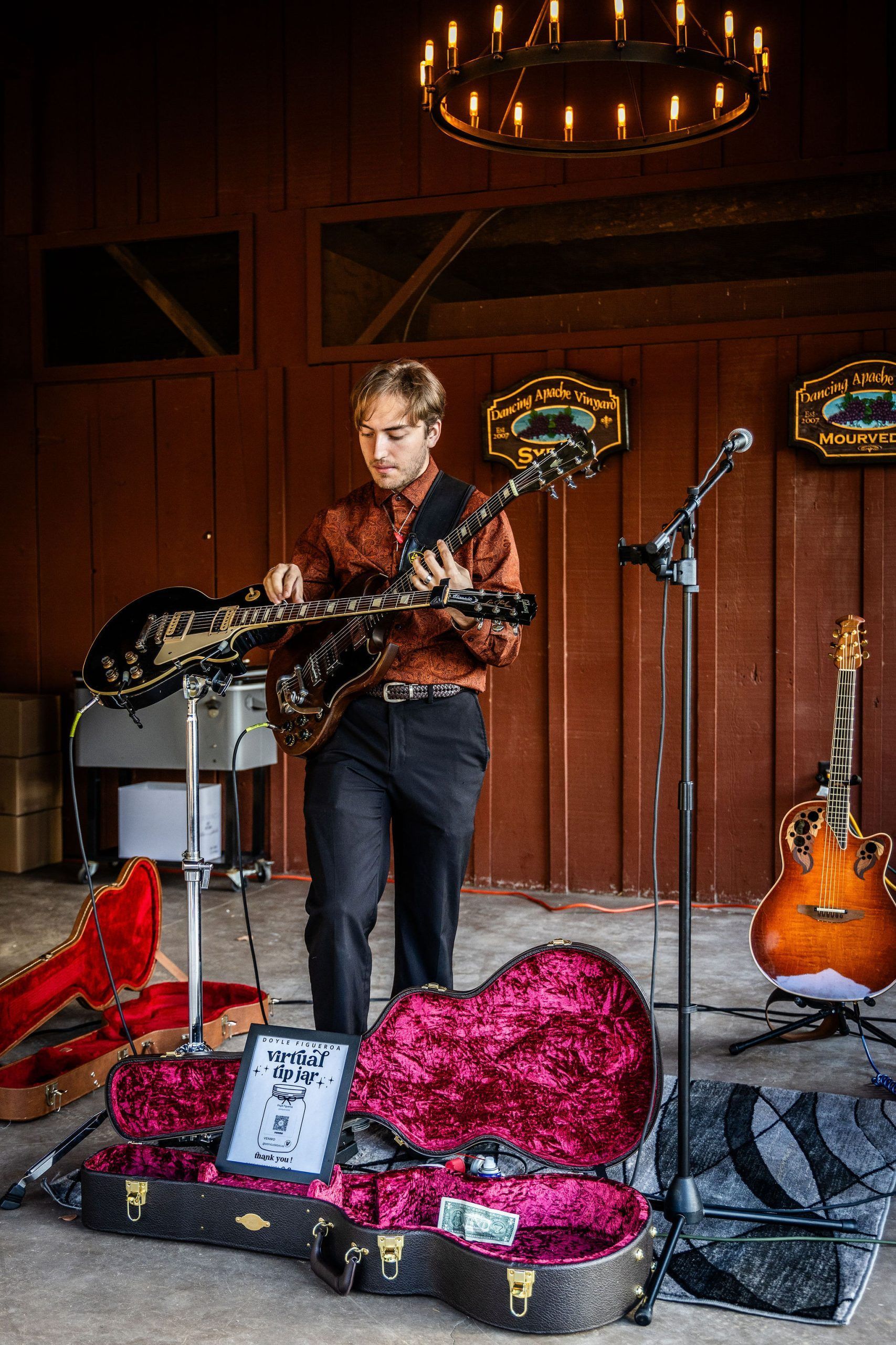 A young man playing a double-necked guitar indoors. He's wearing a patterned shirt and dark pants. Cases and a microphone stand are in view.