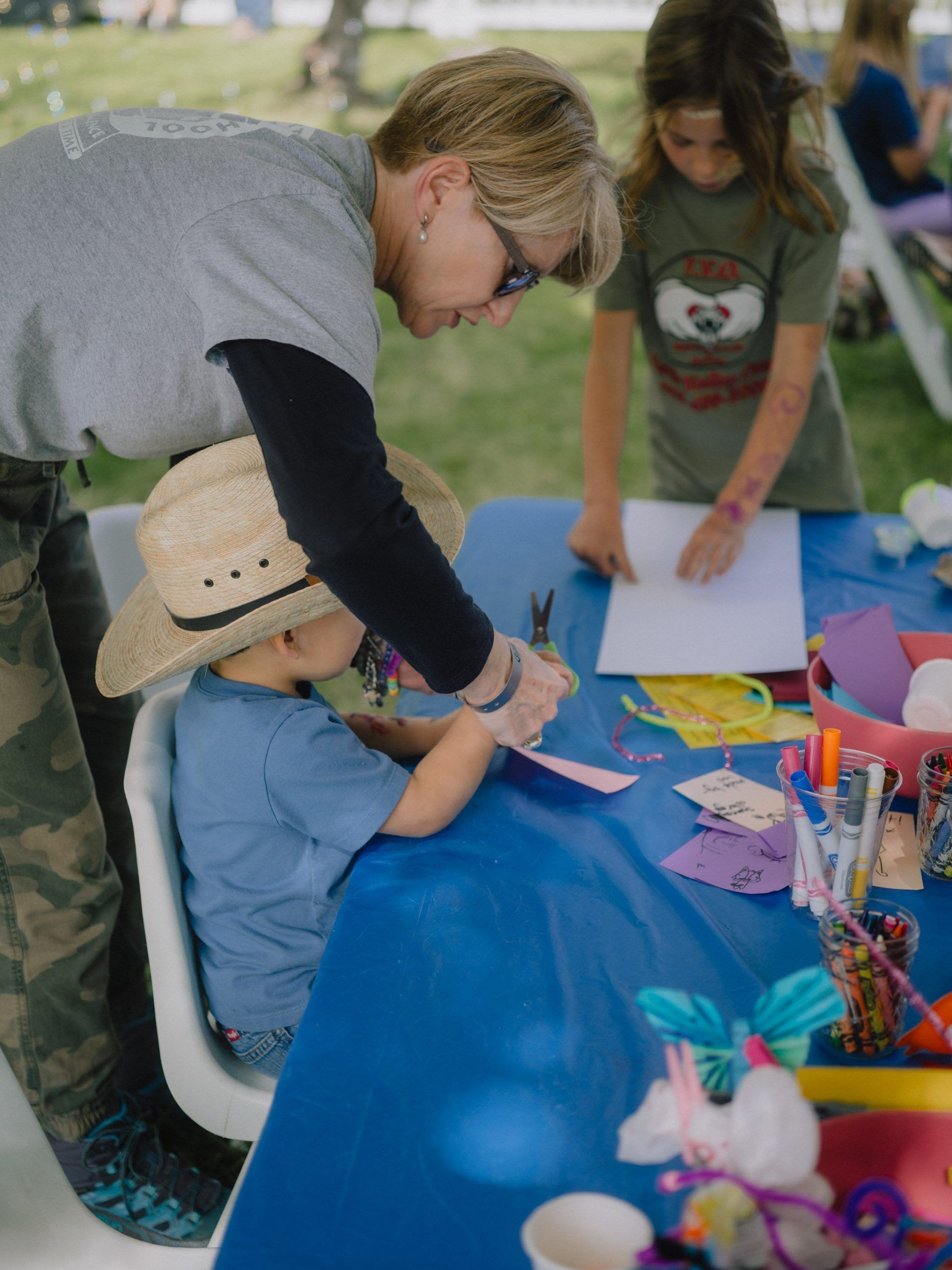 Woman assisting a child with crafts at a blue table; another child looks on. Outdoors, daytime.