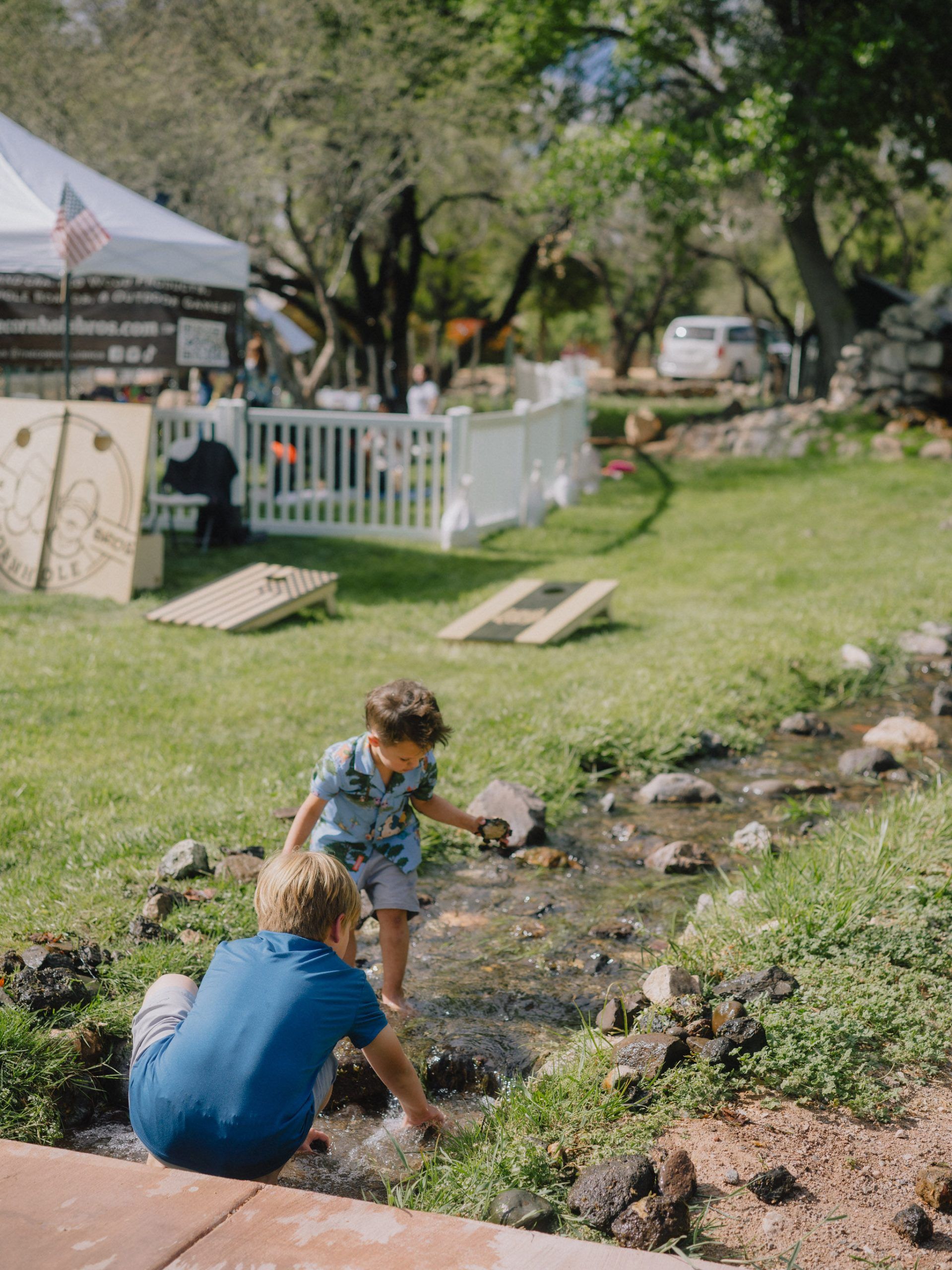 Two children playing in a stream in a park, with a white tent and yard games in the background.