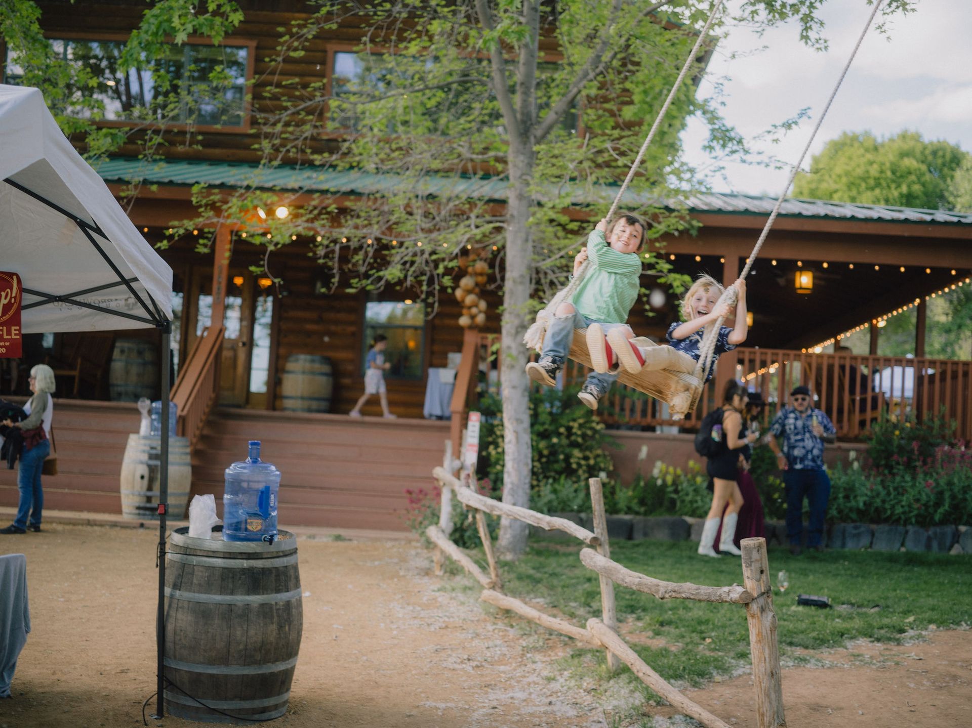 Children on a wooden swing in front of a wooden cabin during a daytime event.