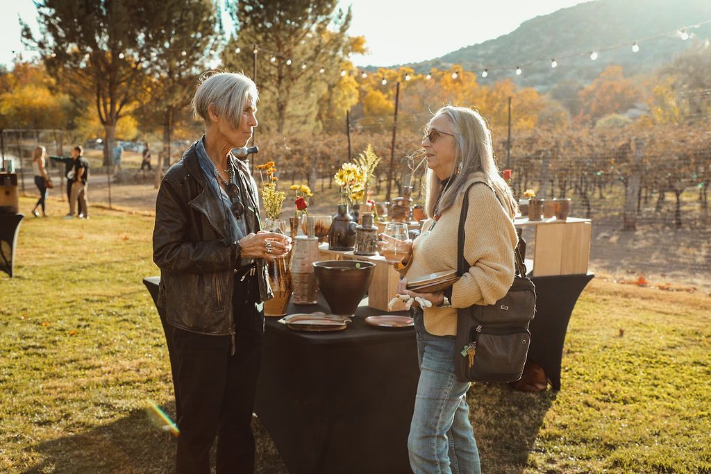 Two women at an outdoor event near a vineyard, conversing by a table with food and drinks.