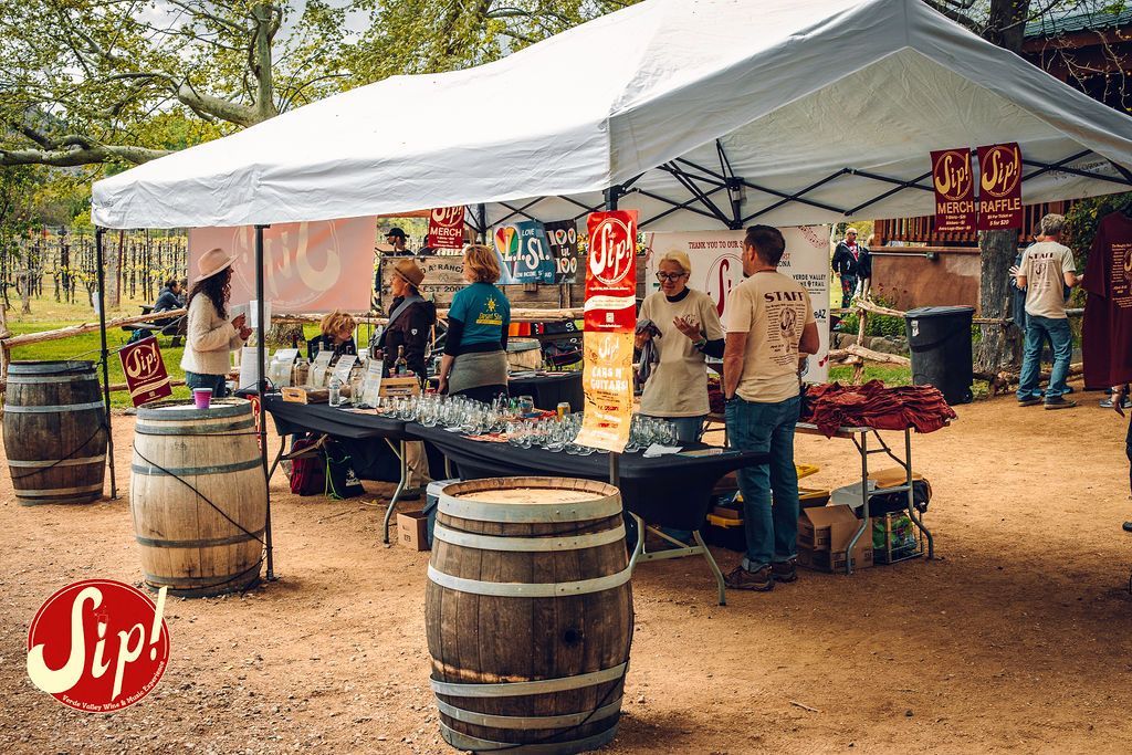 Wine tasting event under a white tent. People serve, taste, and interact at tables with barrels.