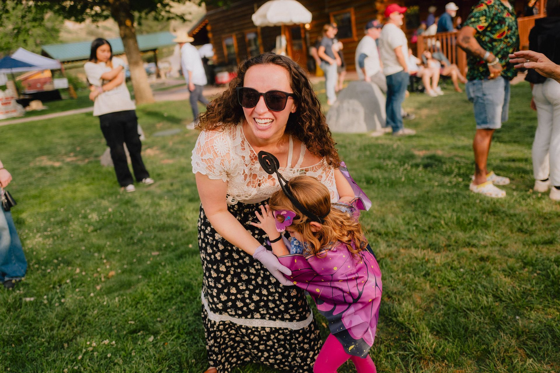 Woman with sunglasses hugs a child at an outdoor gathering; both laugh, surrounded by others.