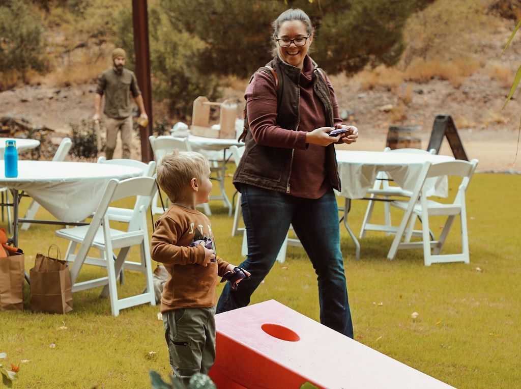Woman smiles, throws beanbag at red cornhole board, child watches. Outdoor party with white tables and a man.