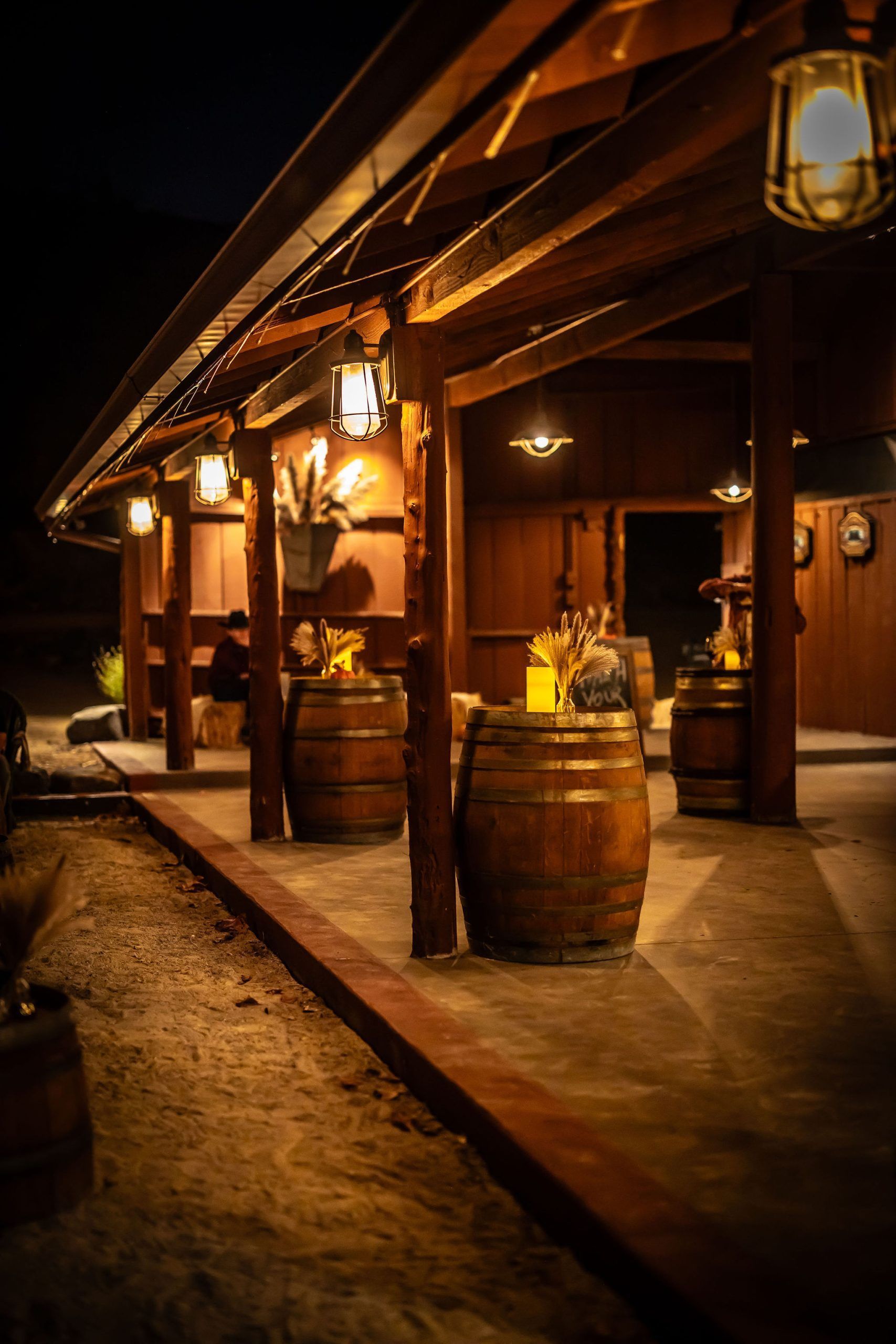 Outdoor evening scene at a winery: wooden structure with barrels, hanging lights, and dark background.