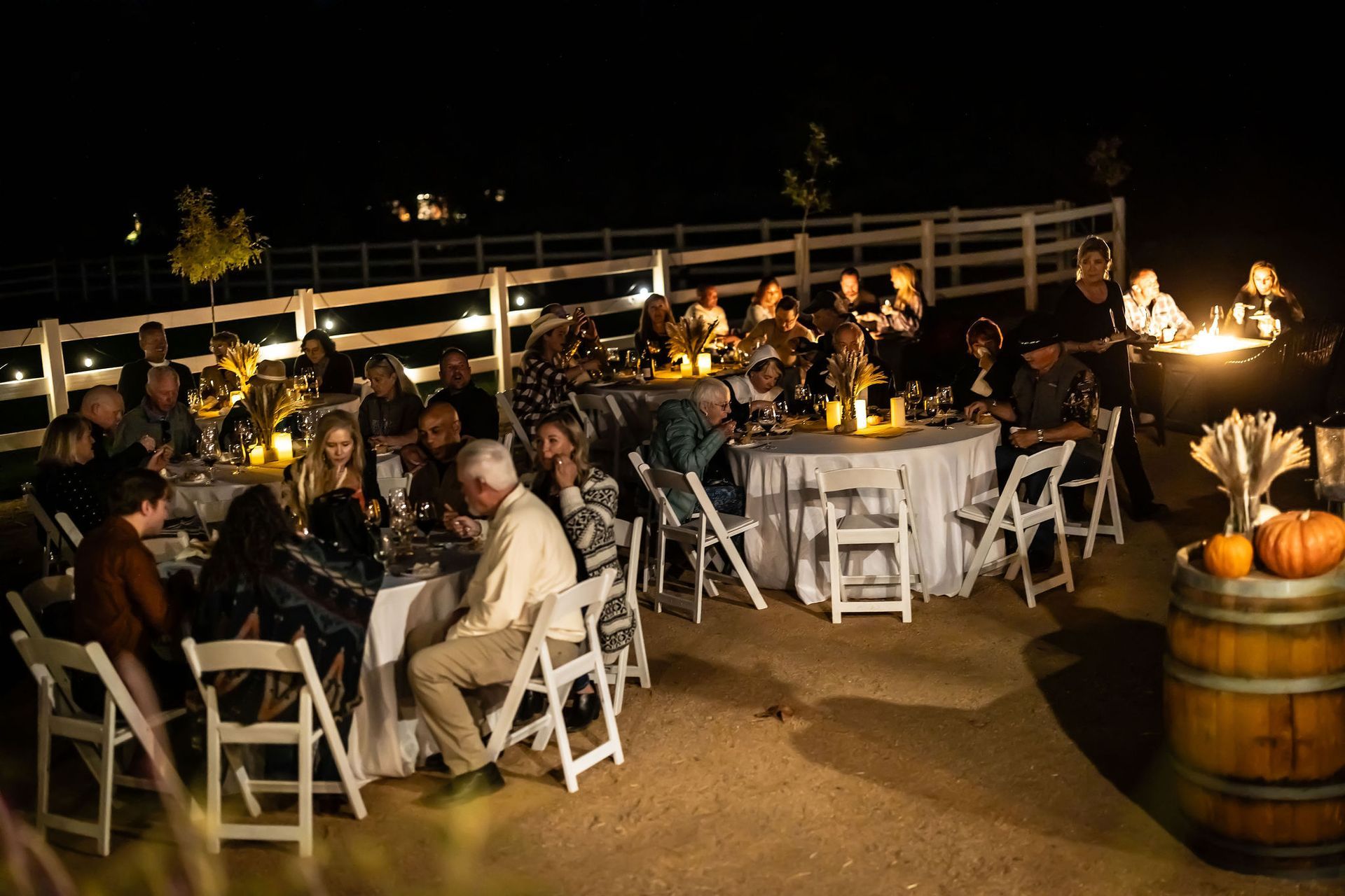 Outdoor nighttime dinner party with guests seated at tables, illuminated by candles and a fire pit.