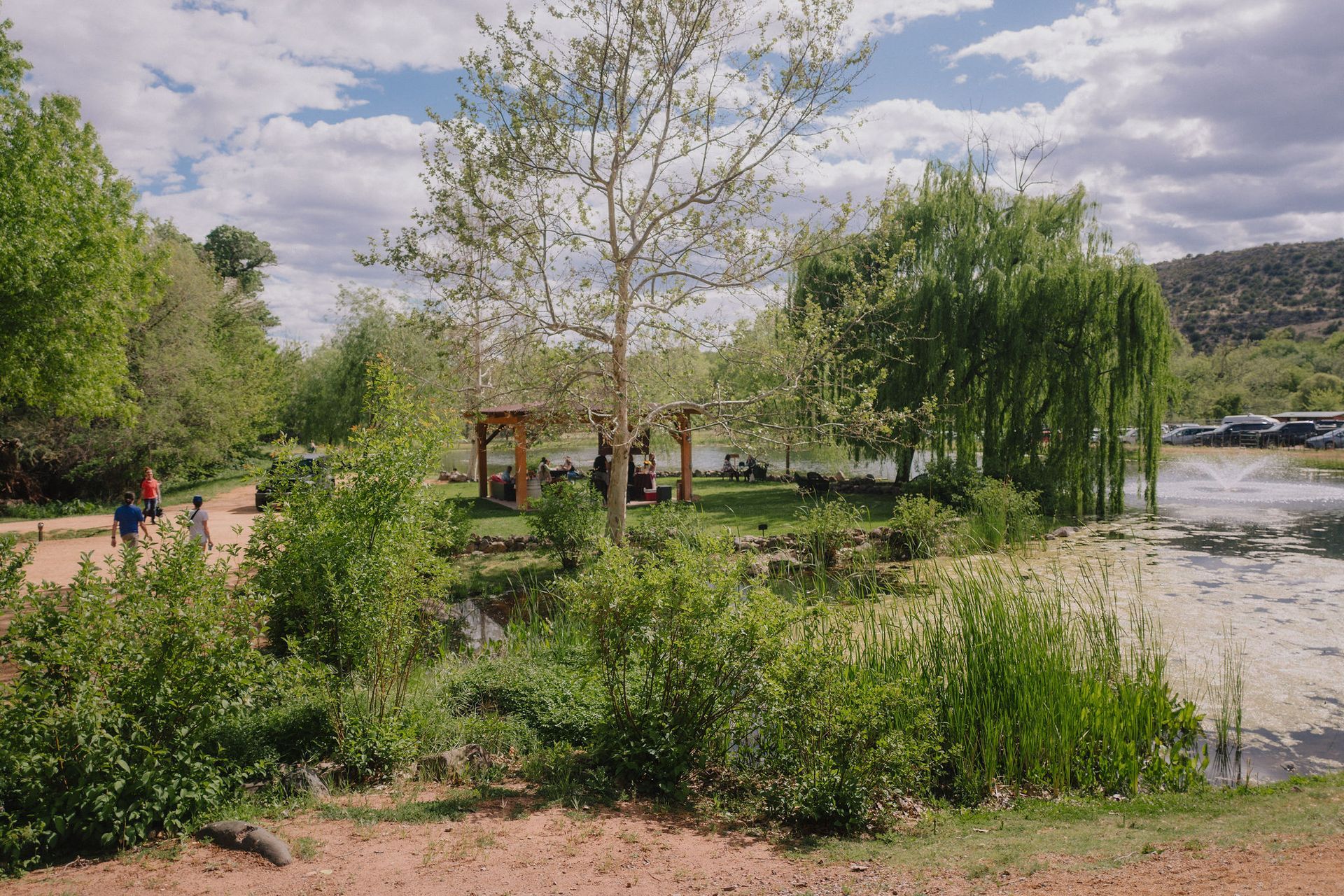 Lush green park with a lake. A gazebo sits near the water. People walking on a path. Overcast sky.