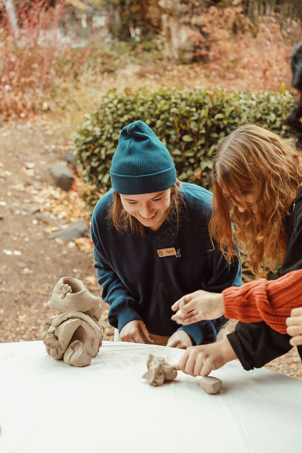 Two people sculpt clay at a table outdoors; one wears a blue hat and smiles.