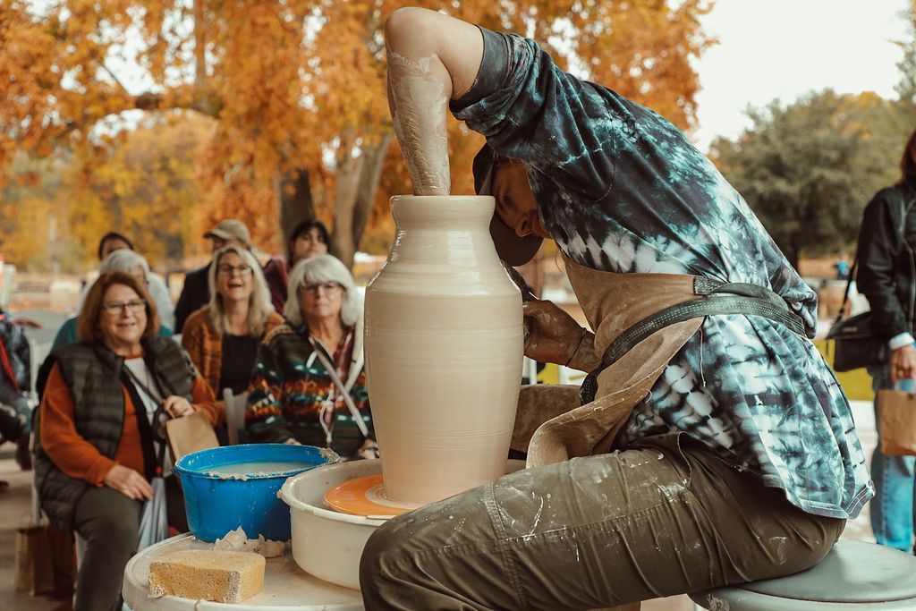 A potter throws a vase, outside. People watch as he works with clay.