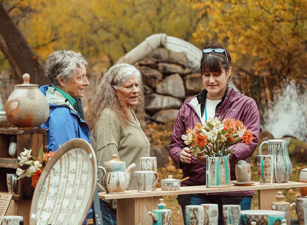 Three women looking at pottery displayed on a table at an outdoor market. Autumn foliage in background.