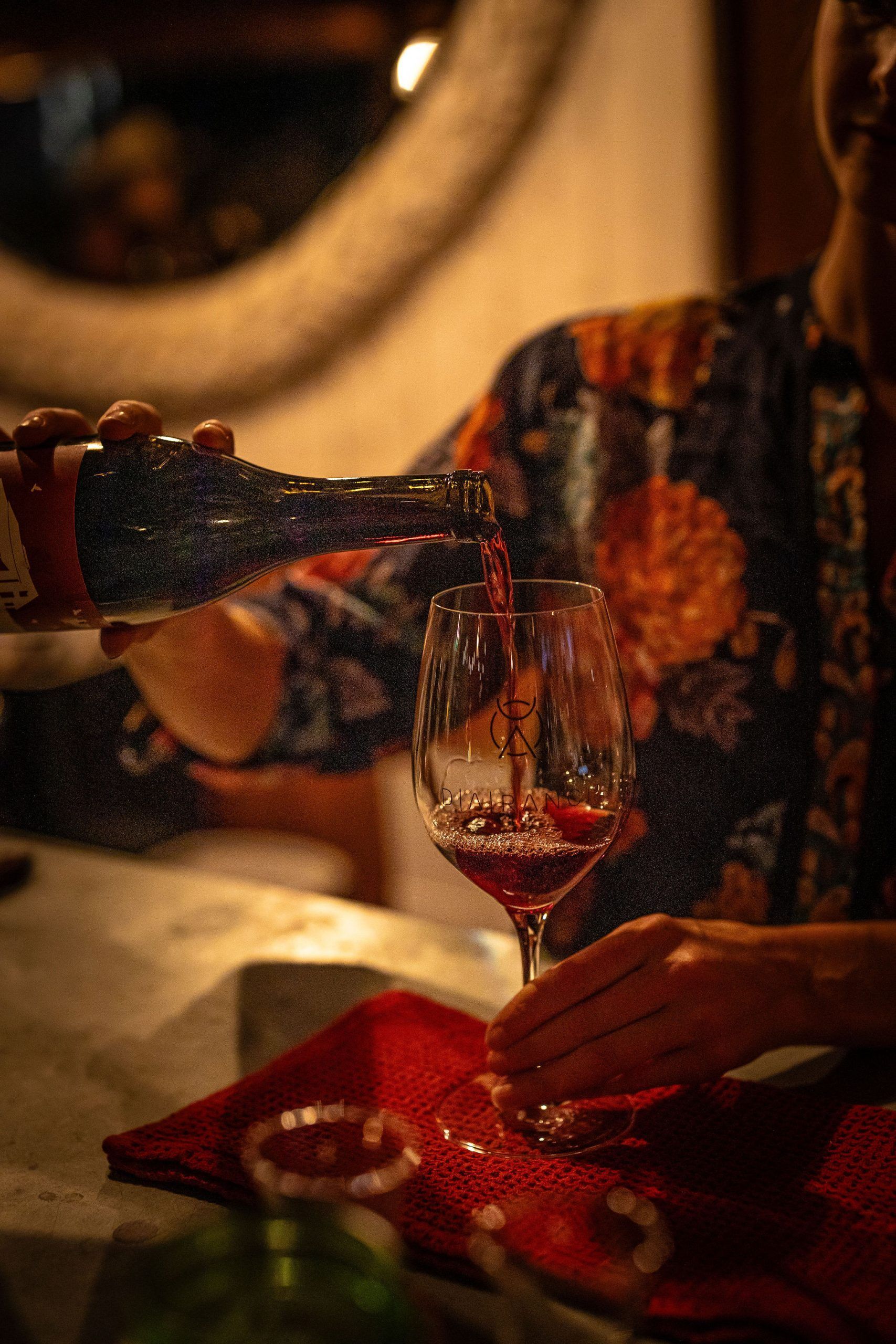 Person pouring red wine into a glass; dark setting, floral shirt.