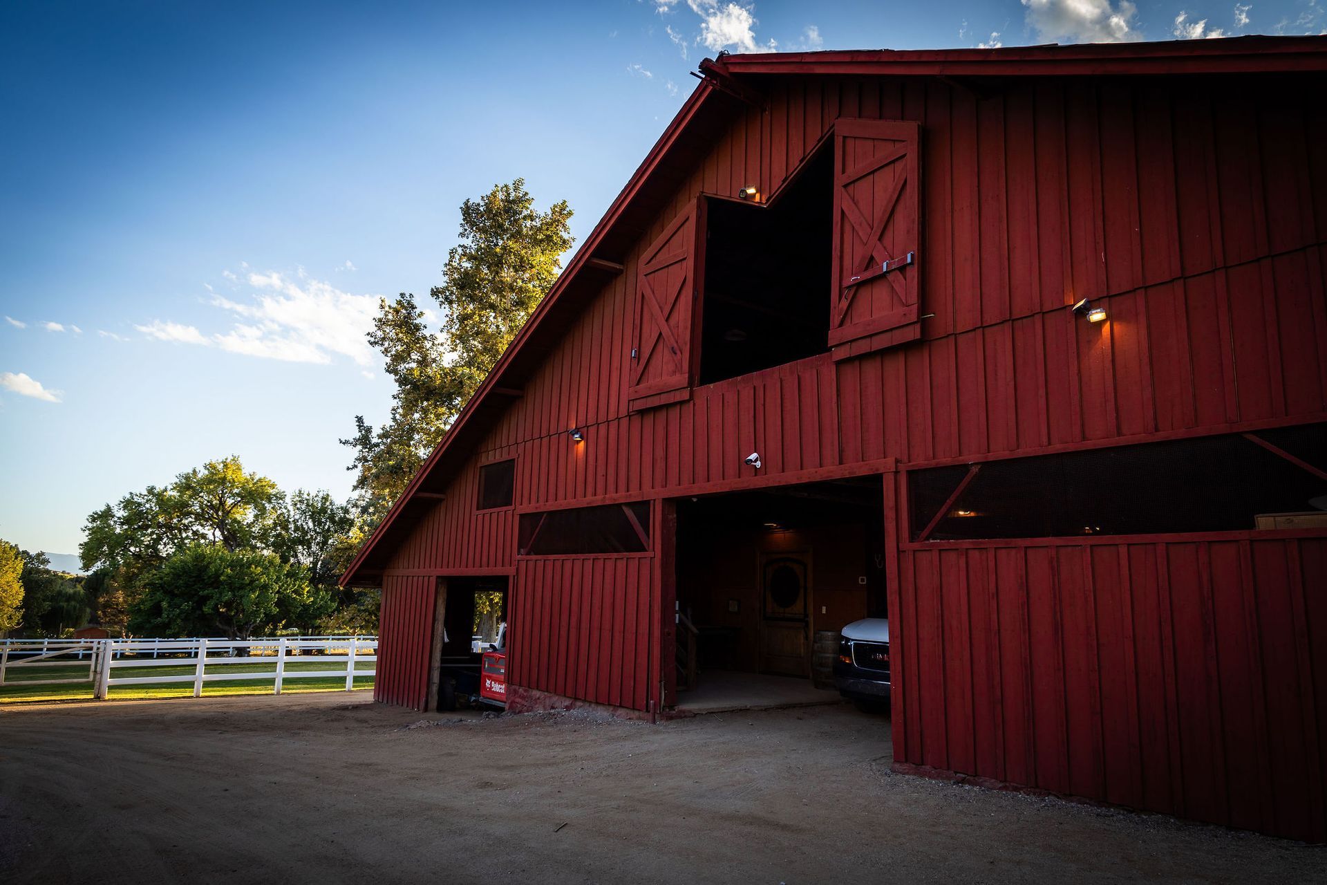 Red barn with open doors and shutters, white fence, trees, and blue sky.