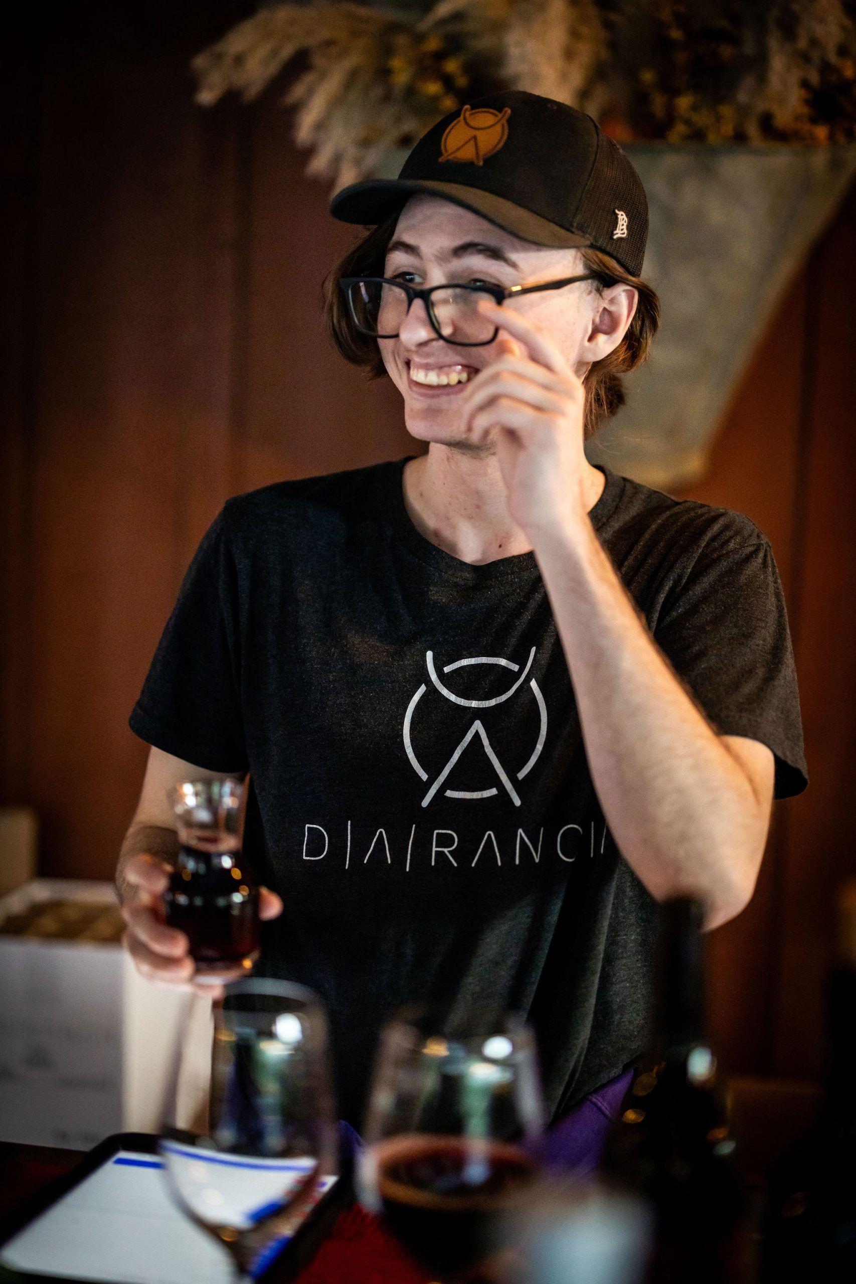 Person smiles, holding wine glass, wearing a black cap and t-shirt, at a winery with glasses on a table.