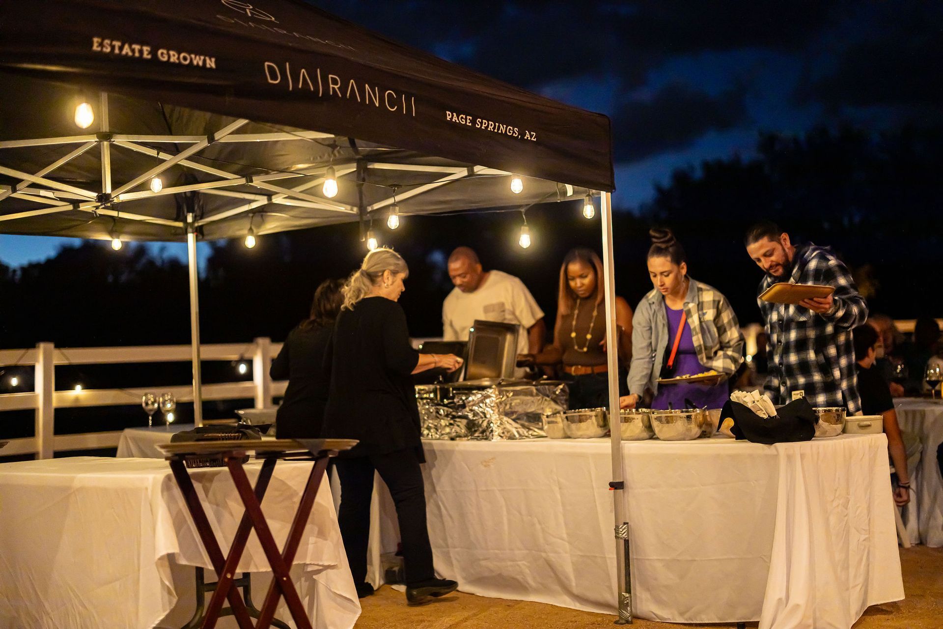 People at a buffet under a tent at night. String lights illuminate food tables.