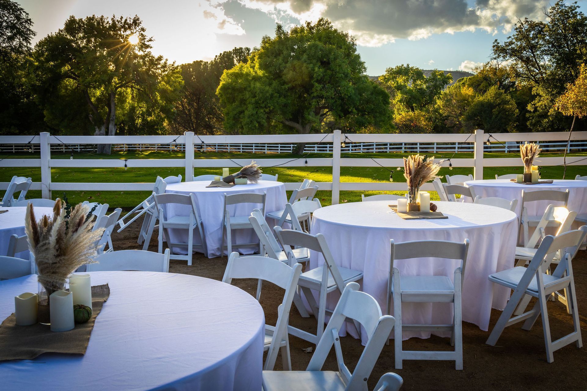 Outdoor event with round tables, white chairs, and decorations. Trees and white fence in background.