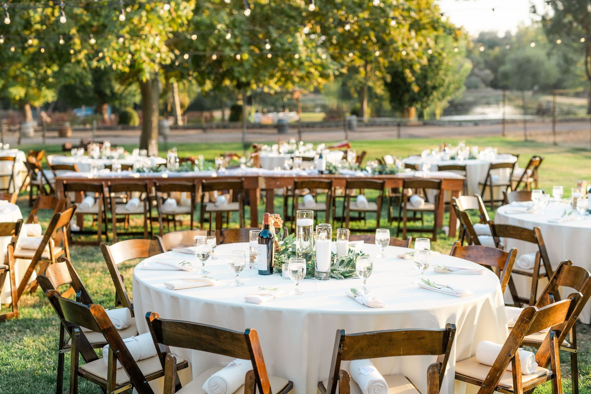 Outdoor wedding reception with round and rectangular tables, wooden chairs, and string lights.