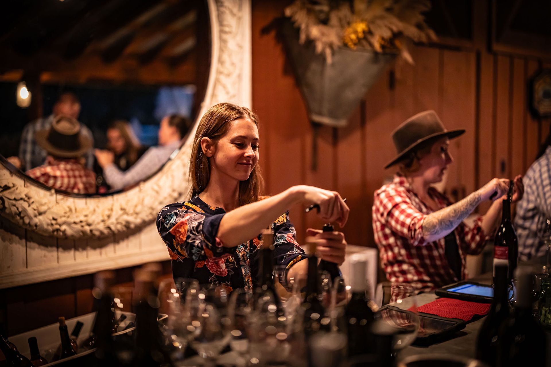 Woman opening a wine bottle at a bar. Another person opens one next to her. Dim, rustic setting.