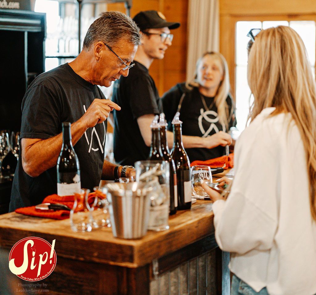 People at a wine tasting: man in black shirt points, others chat, wine bottles and glasses on bar.