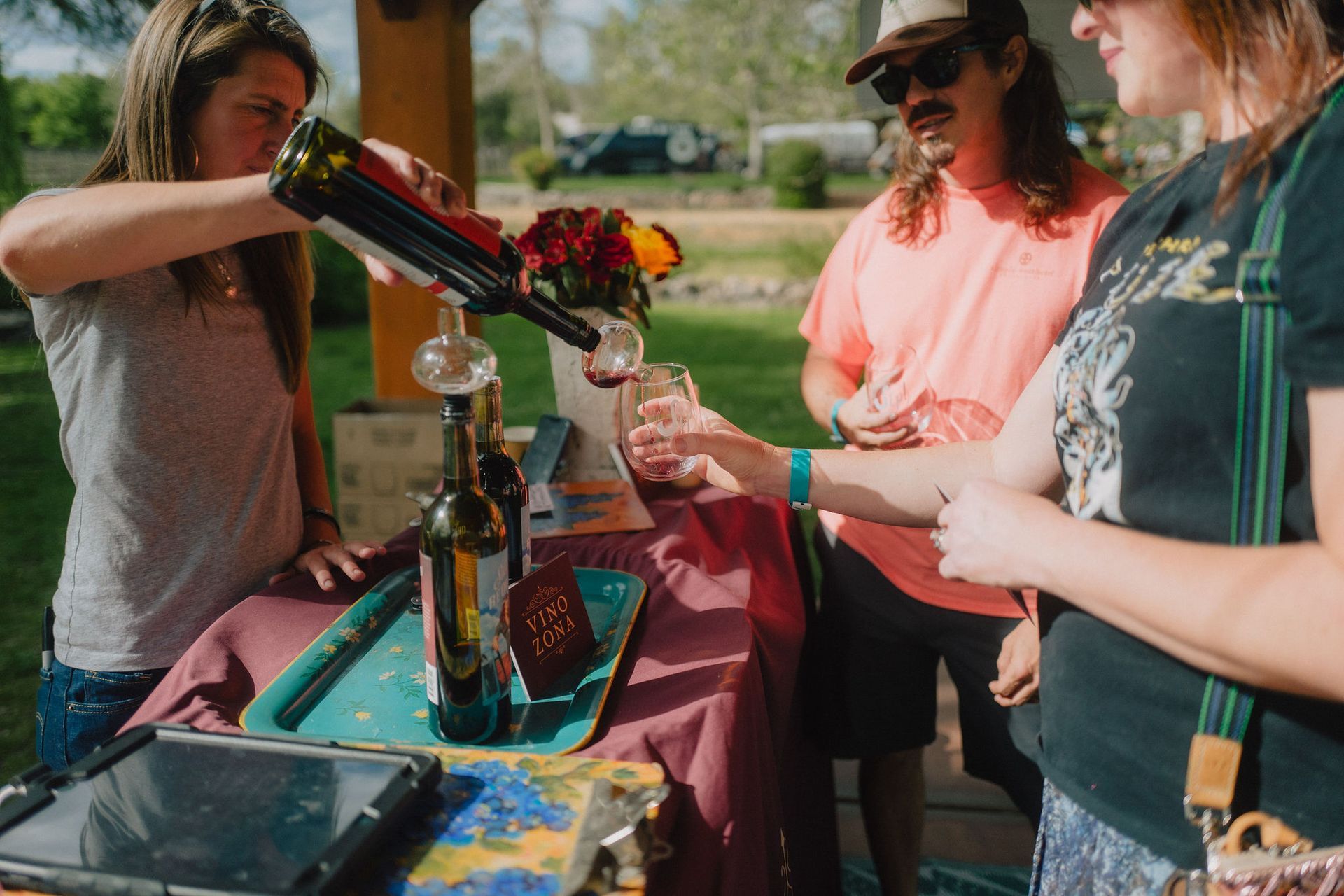 Woman pours red wine at an outdoor wine tasting. Other people watch. Bottles and flowers on a table.