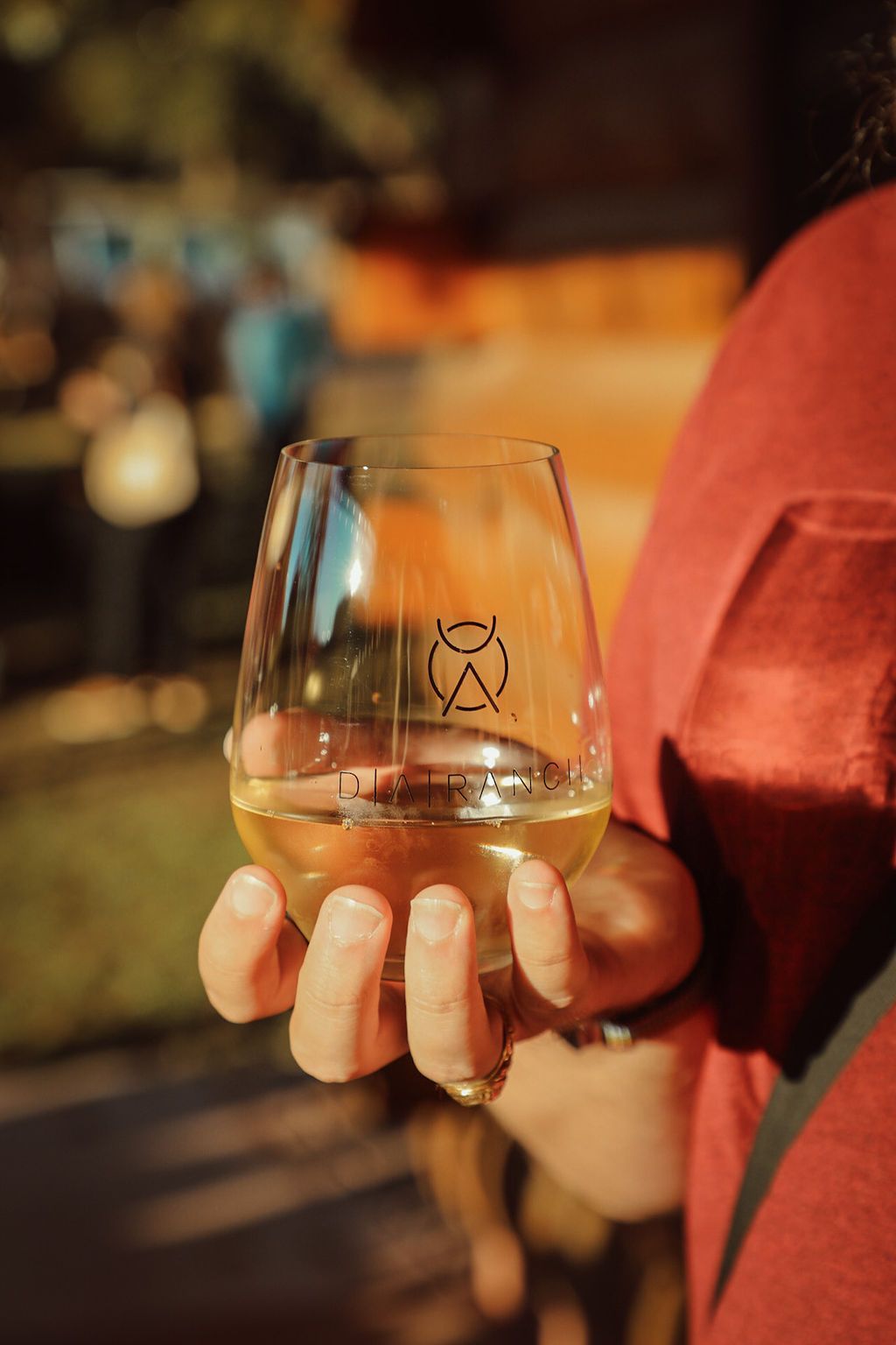 Person holding a wine glass with amber liquid, outdoors. The logo is visible on the glass.