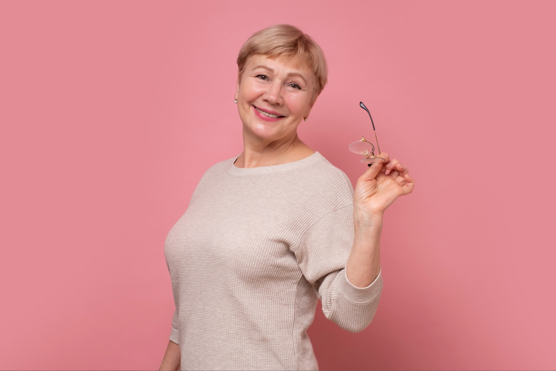 Smiling woman in a beige sweater holding glasses against a pink background