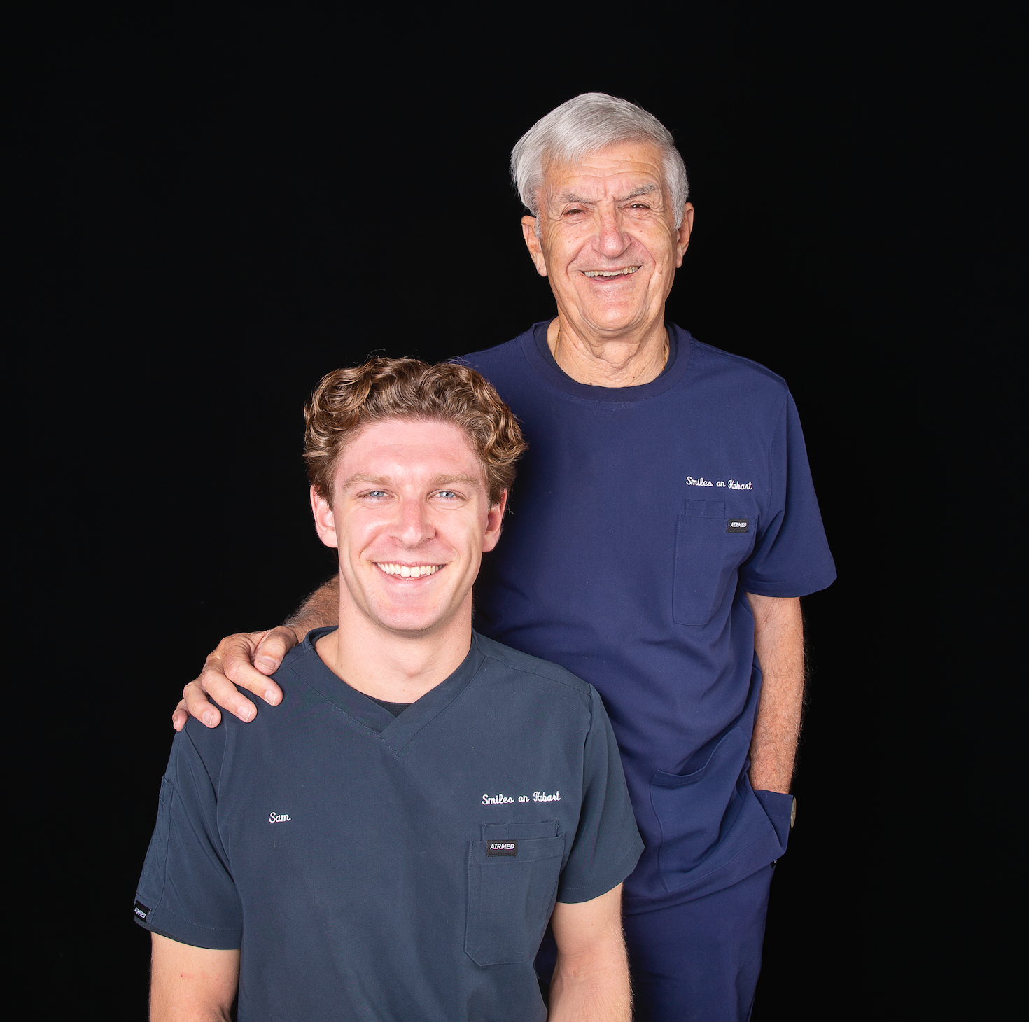 Two smiling people in navy shirts posing against a black background