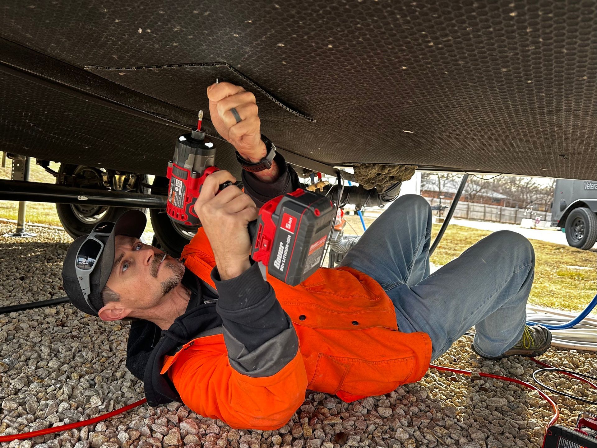 A man in an orange jacket uses power tools to work under a vehicle. Gravel ground with black undercarriage.