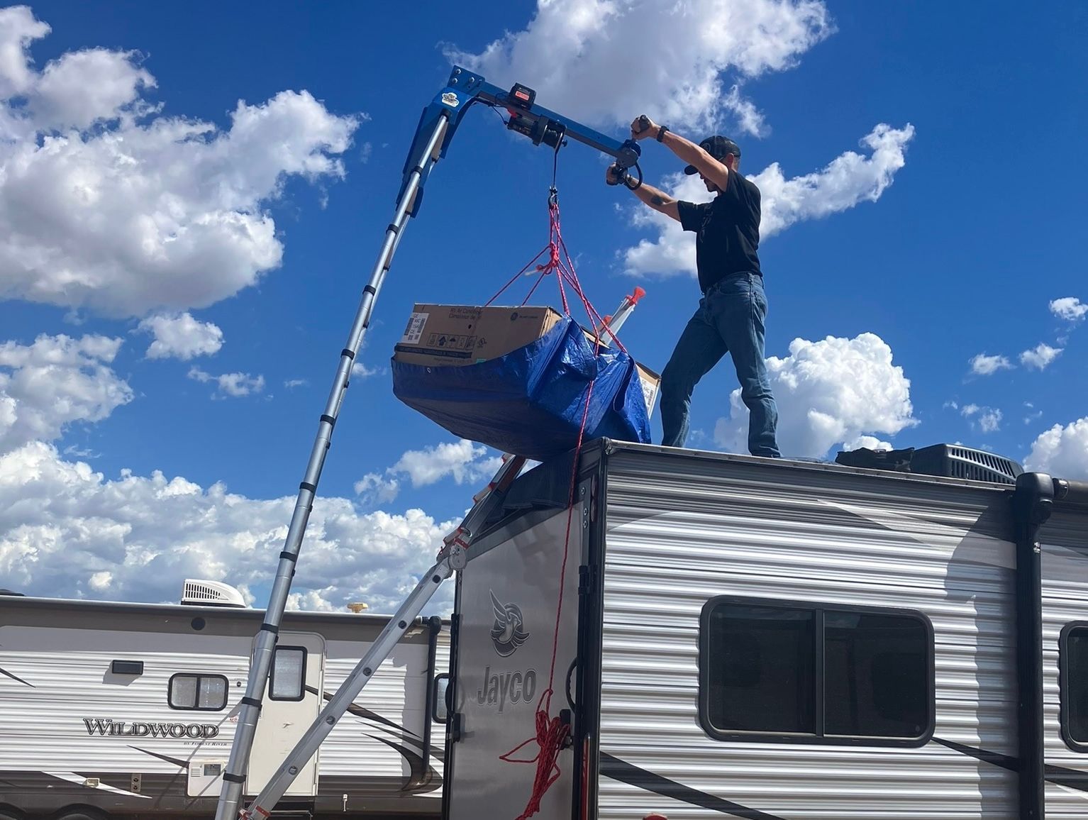 Man using a hoist to lift a blue object onto an RV roof under a cloudy sky.