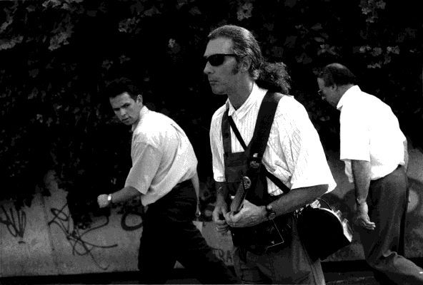 Three men walking outdoors, one wearing sunglasses, a portable amplifier and an electric guitar.
