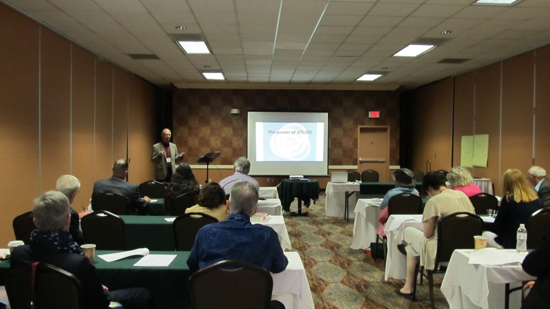 A presenter speaks to a seated audience in a conference room with a screen displaying a circular diagram.