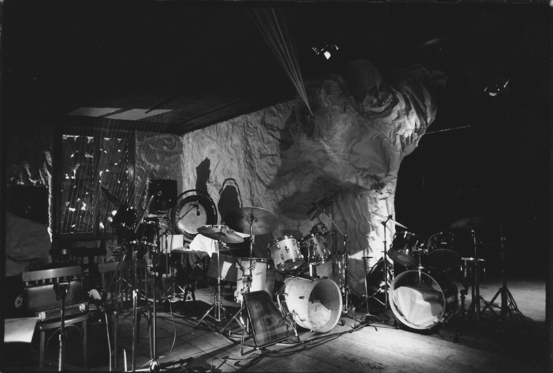 Black and white photo of a drum kit on a stage with a large face in the background. Spotlights shine.
