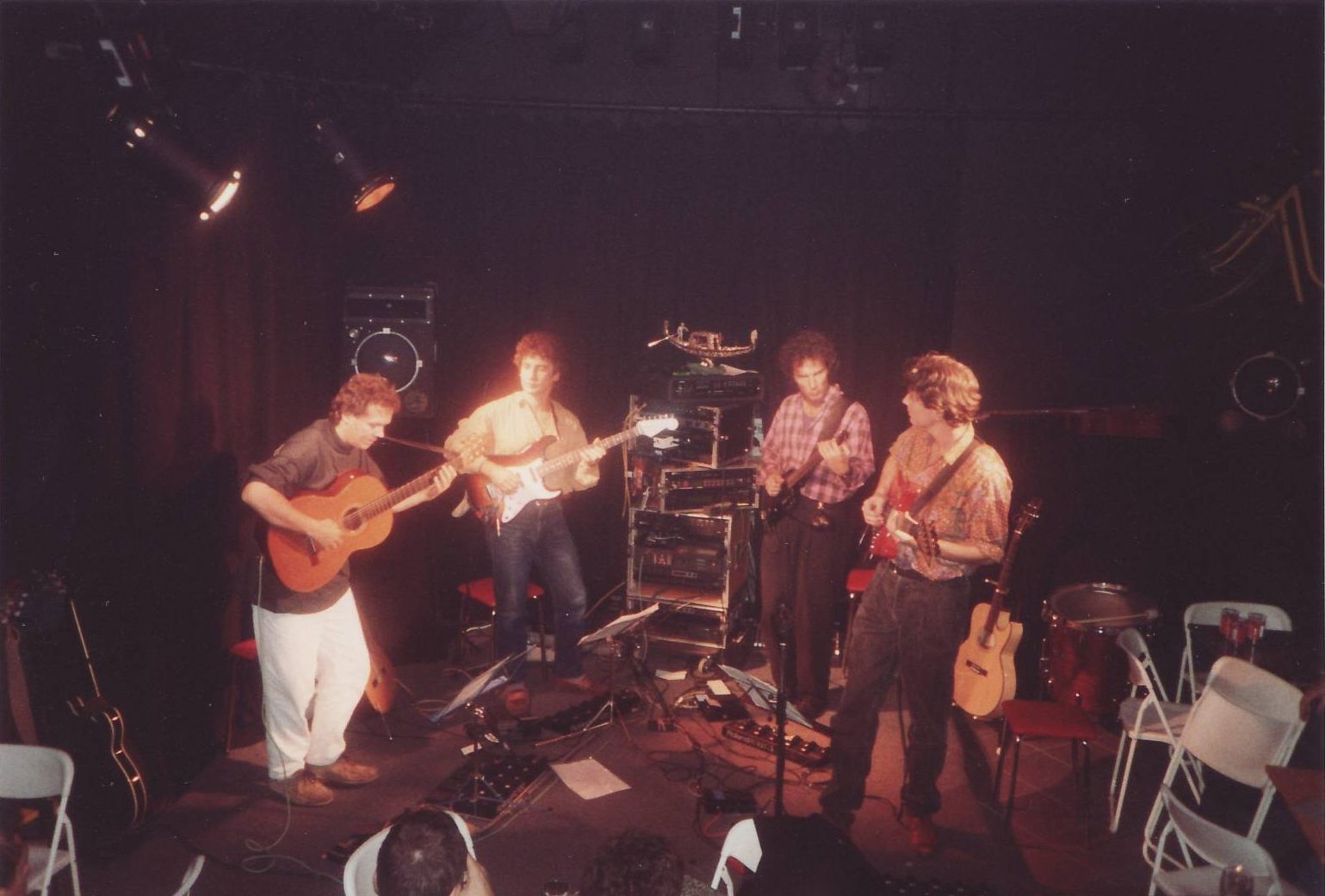 Four musicians on a dimly lit stage playing instruments: acoustic guitar, electric guitar, and percussion.